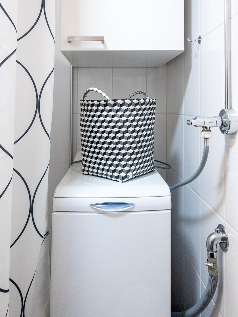 A compact laundry area features a washing machine topped with a patterned storage basket. The surrounding space is tiled in a light color, complemented by a shower curtain with a simple geometric design. Wall-mounted storage adds functionality to the design.