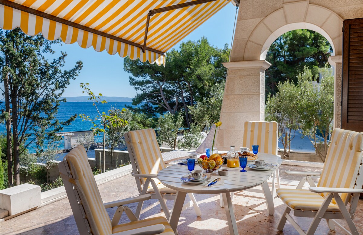 A covered outdoor patio features a round dining table set with blue glasses, plates, and a fruit bowl. Sun loungers with white and yellow stripes provide seating. In the background, the sea view is framed by lush greenery and clear skies.