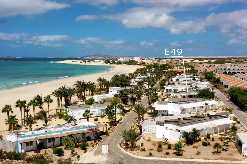 This aerial view captures a coastal landscape featuring a stretch of sandy beach alongside palm trees. The area is dotted with white buildings, with roads winding through the greenery, leading toward the ocean and distant dunes under a partly cloudy sky.