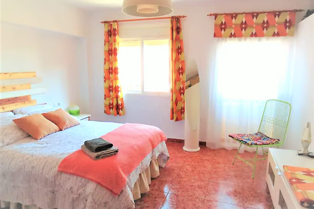 A bright bedroom features a spacious bed adorned with soft linens and decorative pillows. Large windows allow natural light, while colorful curtains and a green accent chair add vibrancy. A bedside table with a lamp complements the inviting atmosphere.