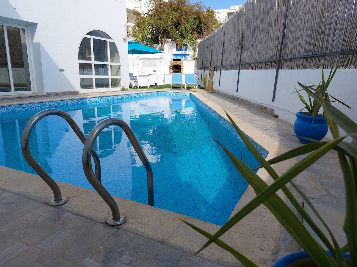 A glistening swimming pool is framed by a smooth stone deck, with metallic handrails providing access. Potted plants are positioned along the edges, enhancing the serene environment. Light reflects off the clear water, creating a vibrant blue hue throughout the space.