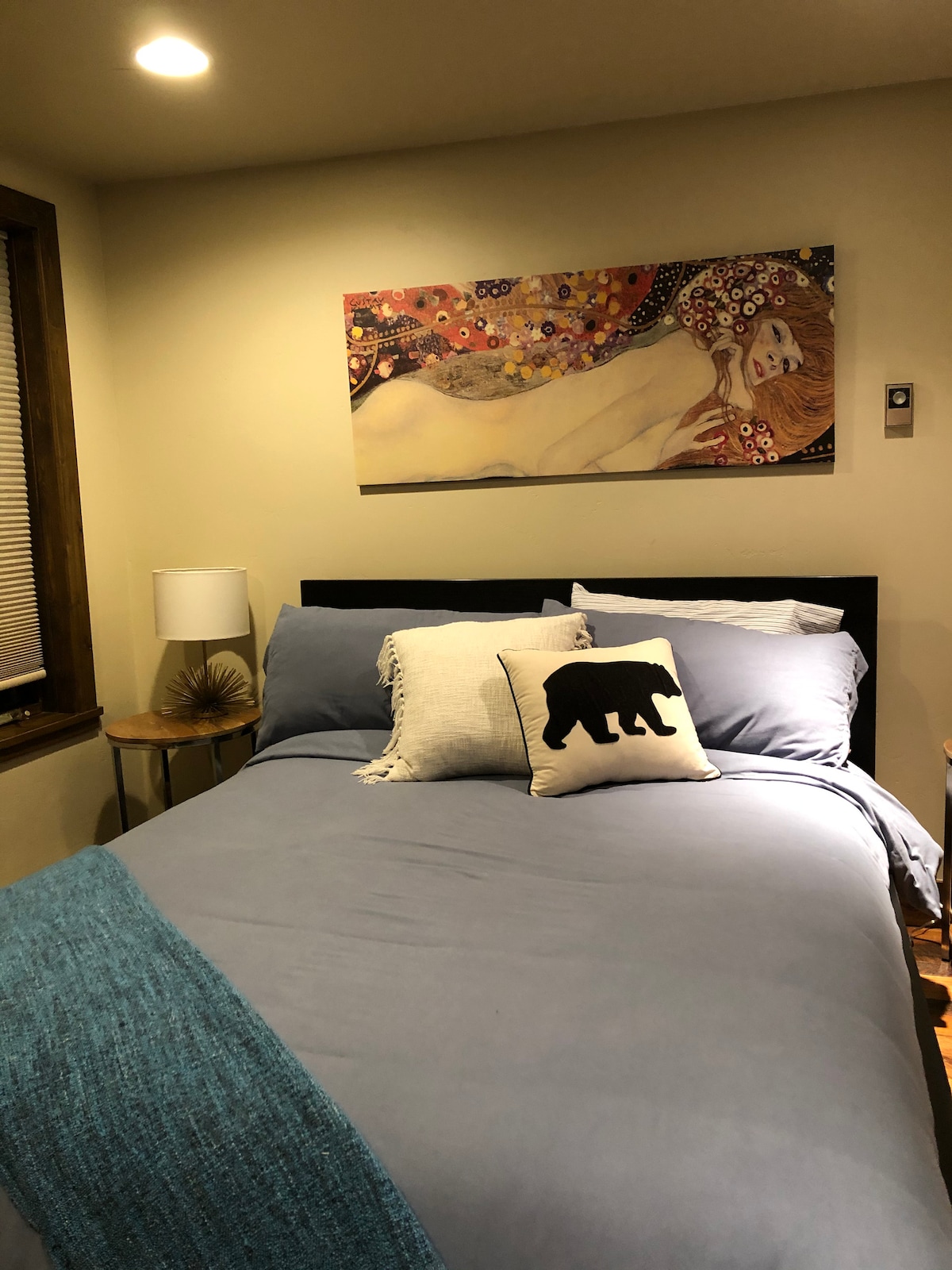 A cozy sleeping area features a spacious bed dressed in blue linens, complemented by decorative pillows. A piece of art adorned with warm colors is displayed above the bed, and a bedside lamp stands next to a small decorative table.