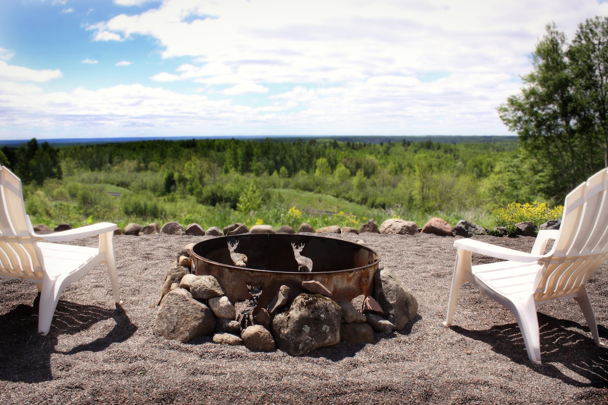 A cozy fire pit surrounded by a circle of stones is positioned on a gravel patio. Two white Adirondack chairs provide seating, while lush greenery and a distant view of the landscape are visible in the background under a partly cloudy sky.