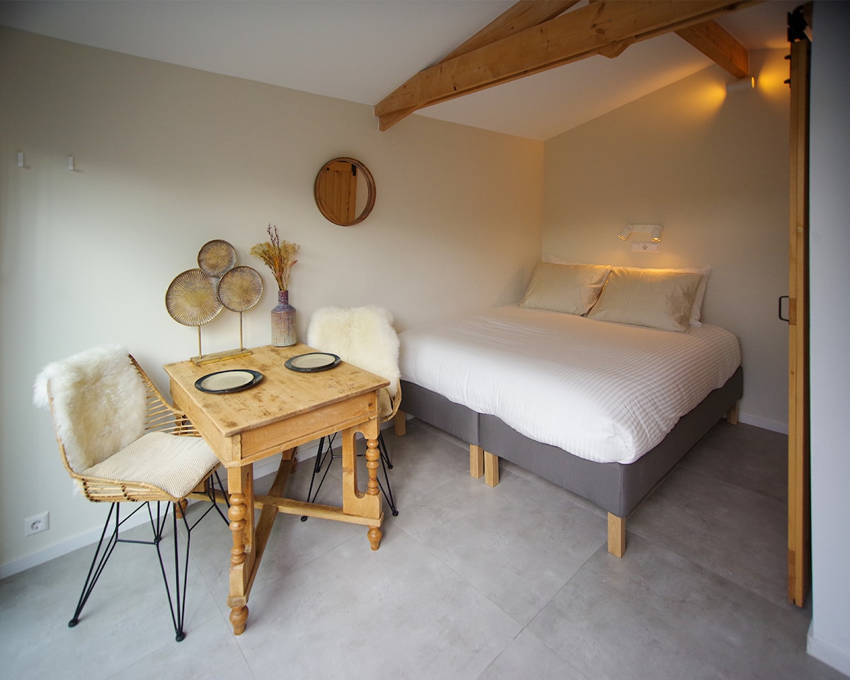 The interior space features a spacious bed with soft bedding, complemented by natural light filtering through a nearby window. A small wooden table is set for two, accompanied by minimalist chairs, while decorative plates and a vase with dried flowers add a subtle touch.