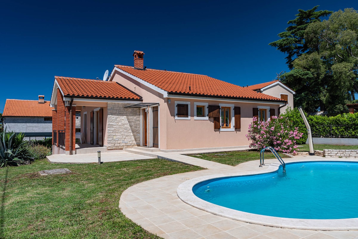 A modern villa is showcased, featuring a warm exterior with a red tiled roof and light-colored walls. The swimming pool is framed by a paved area and green lawn, while flowering shrubs add a touch of nature to the surroundings.