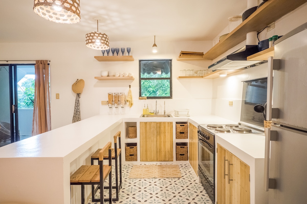 The kitchen features modern appliances and ample counter space, with wooden shelves displaying dishware. A row of stools is positioned at the counter. Light streams in through a window, illuminating the patterned floor tiles and creating a welcoming atmosphere.