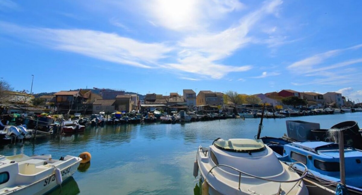 The tranquil waters reflect colorful buildings lining the small port, with boats moored gently along the canal. Bright blue skies are visible, adorned with scattered clouds. The scene captures a serene atmosphere typical of the local fishing village.