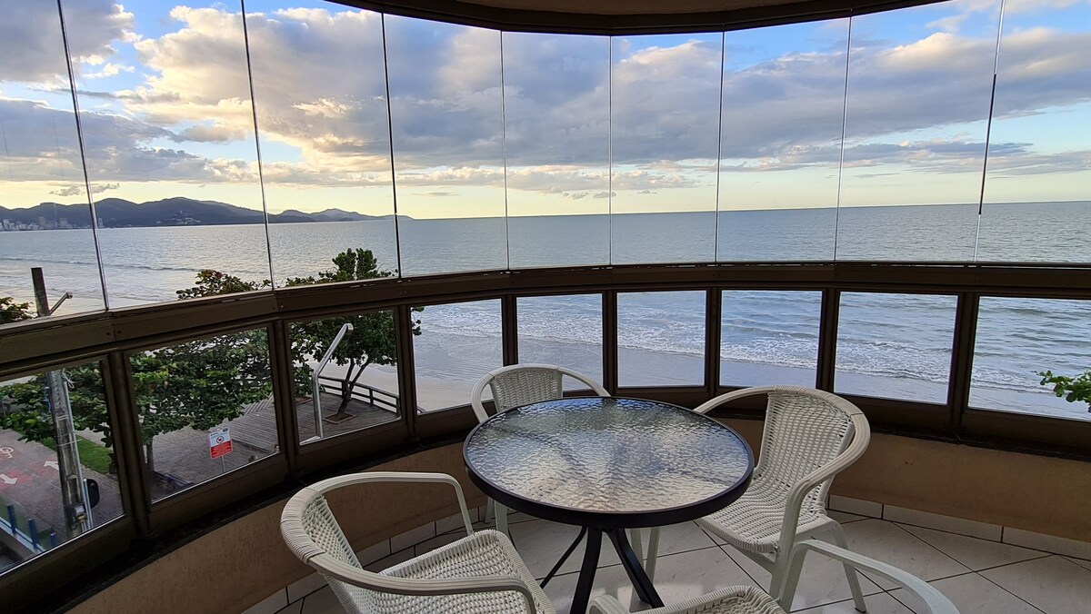 A spacious balcony offers a panoramic view of Itapema beach, featuring large windows that provide unobstructed sightlines. A round glass table is surrounded by four white chairs, inviting relaxation while enjoying the coastal scenery.