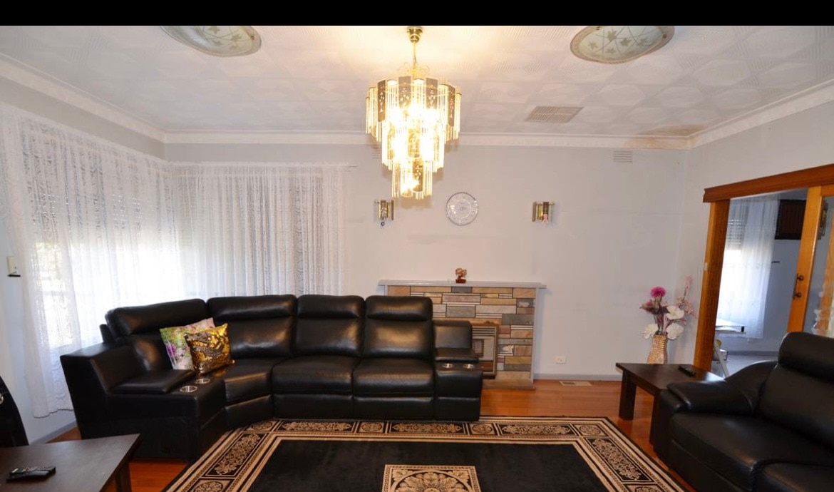 A spacious living area features a sectional black leather sofa arranged around a decorative rug. A chandelier hangs from the ceiling, illuminating the room. Light streams in through sheer white curtains, while a stone fireplace adds a cozy focal point to the space.