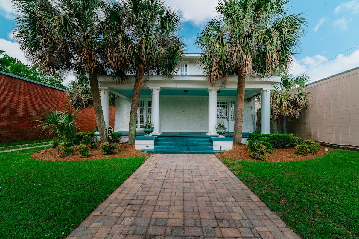 The image captures the exterior of a historic building surrounded by lush greenery and palm trees. A stone pathway leads to the front porch, where white columns support the covered entryway. Fresh landscaping enhances the inviting appearance of the property.
