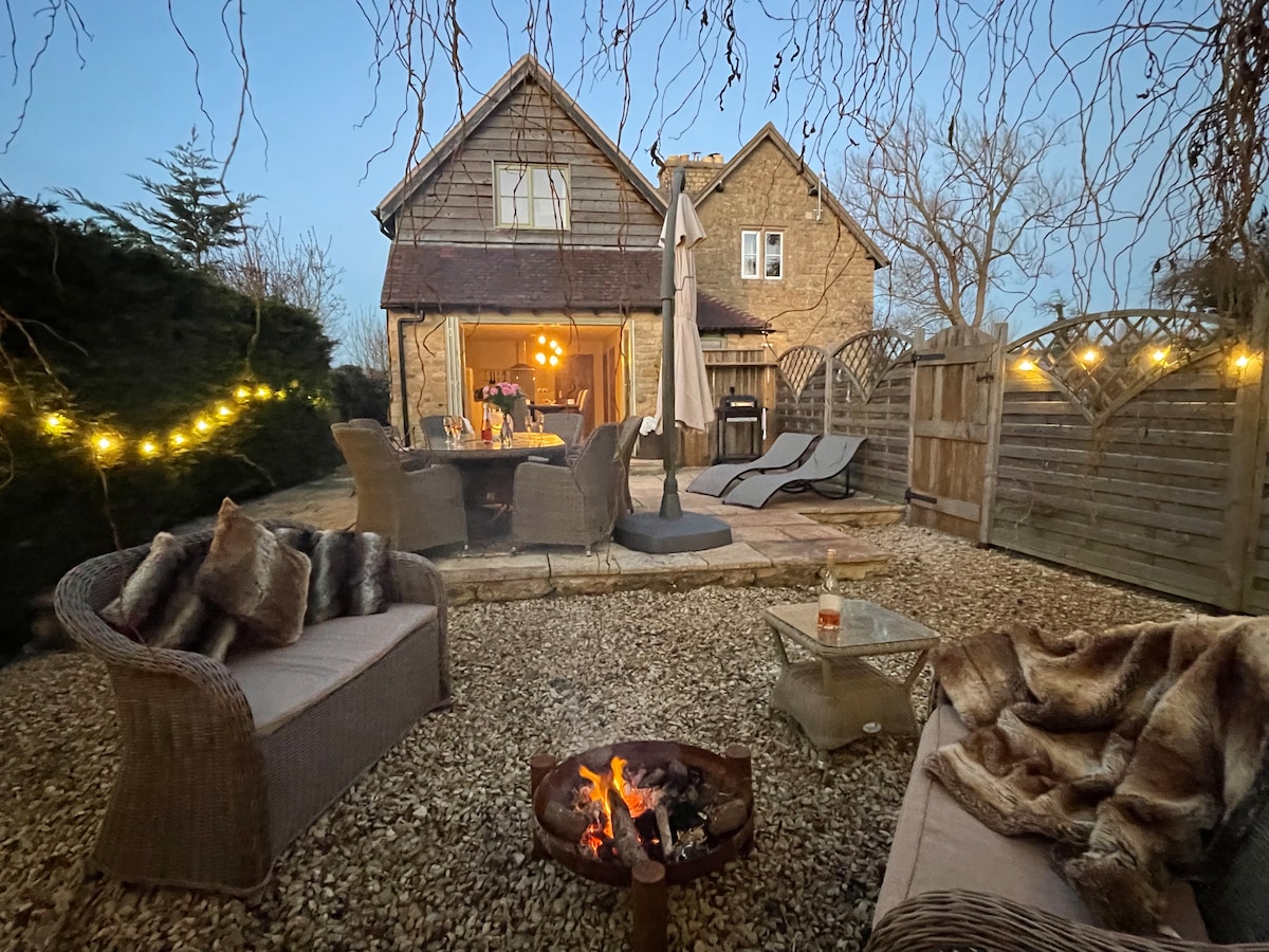 An outdoor seating area features woven furniture arranged around a fire pit, providing a cozy gathering spot. Soft lighting from hanging bulbs creates a warm ambiance, with a dining table set for guests. Sun loungers are visible, complemented by natural greenery in the background.