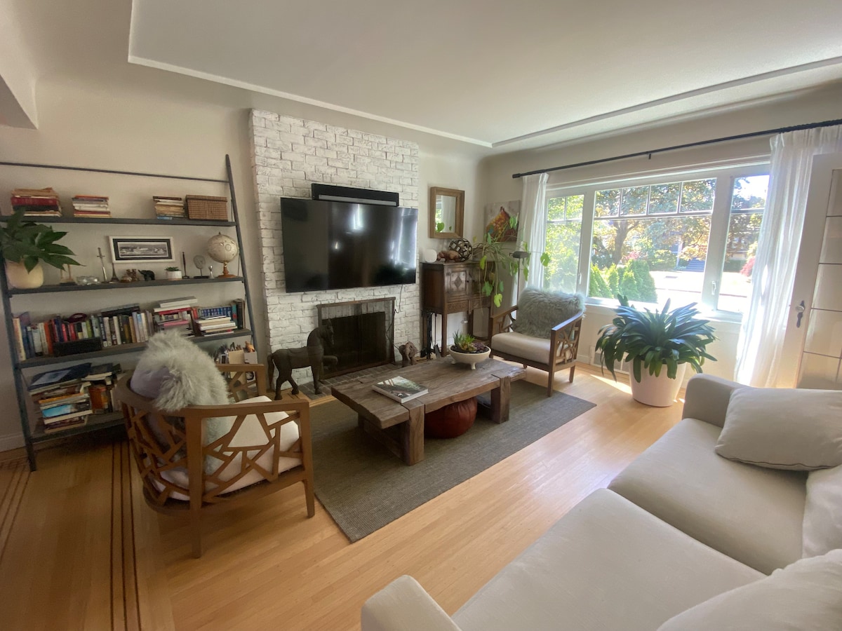 An open living area features a working fireplace set within a white brick wall. Seating includes two armchairs and a sofa, with a coffee table at the center. Natural light floods the space through large windows, revealing views of the outdoors. A bookshelf and indoor plants add character.