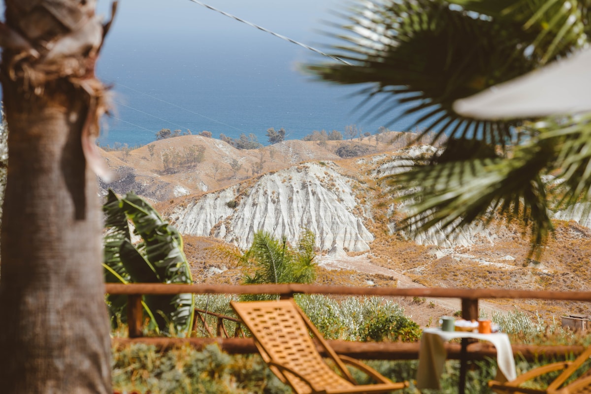 The image captures a scenic view overlooking the coastline, with rolling hills in the foreground and the blue expanse of the Ionian Sea in the background. A lounge chair and a small table are positioned in the garden, framed by lush greenery and palm trees.