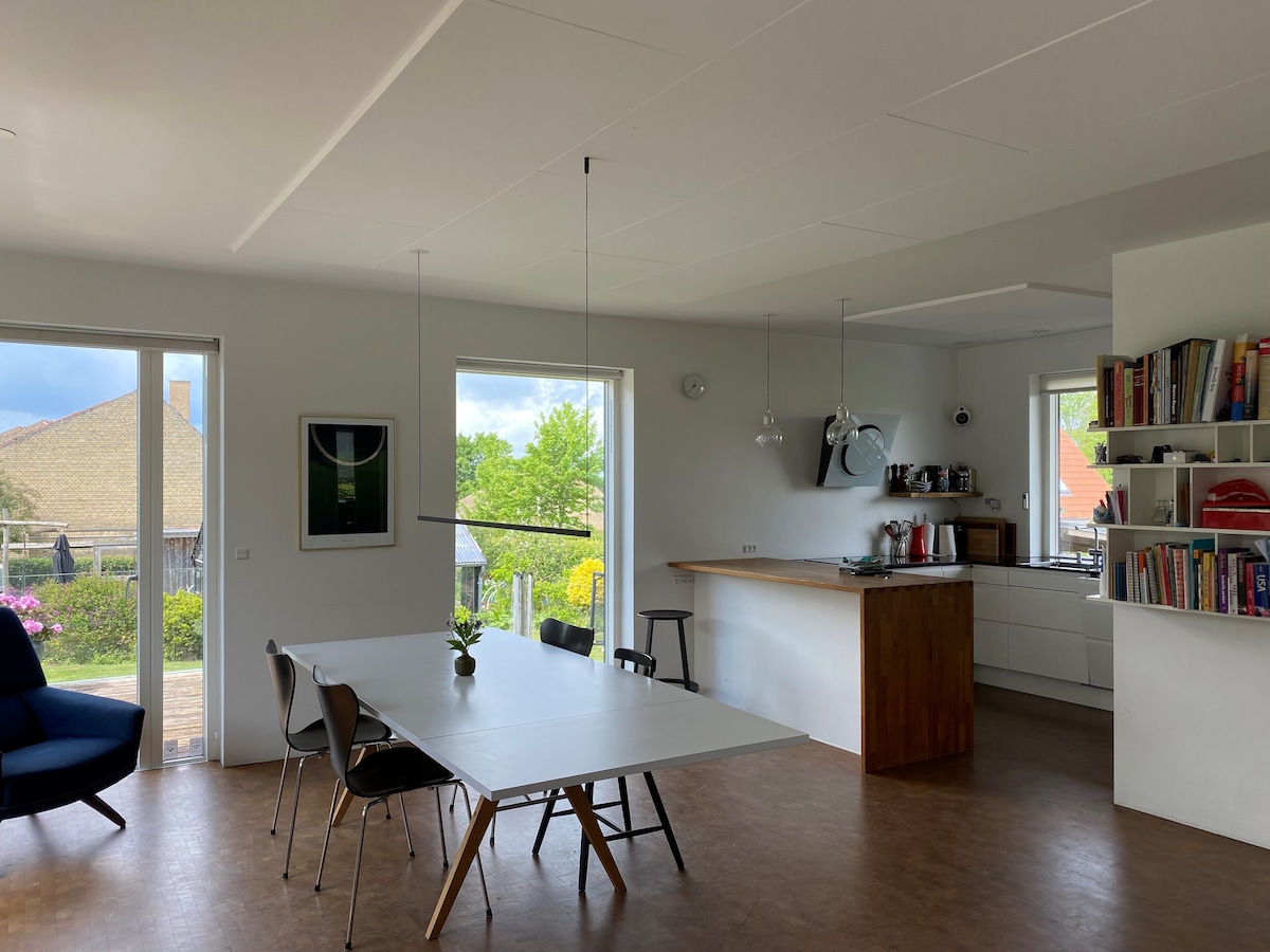 An open-concept living area features a dining table surrounded by simple chairs. Large windows allow natural light, offering views of the garden. A kitchenette is visible, equipped with modern appliances, and a cozy seating area presents a comfortable armchair.