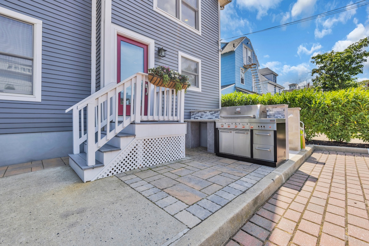 A well-equipped outdoor grilling area is featured beside the private entrance. A stainless steel barbecue grill is positioned adjacent to a small tiled patio. Planters with flowers accent the entryway, complemented by the blue exterior of the home and a neatly arranged hedge in the background.
