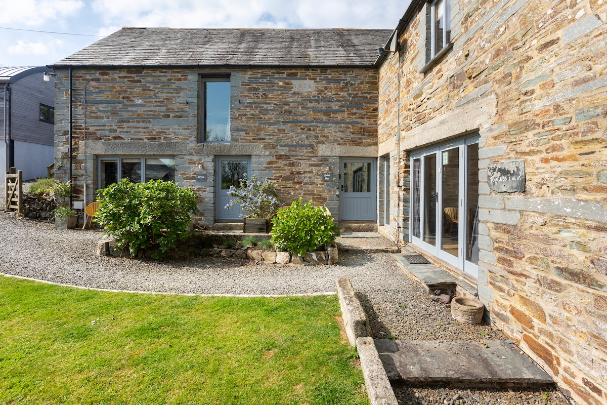 The exterior of Skyber Barn features traditional Cornish stone walls, surrounded by well-tended greenery and a gravel path. Large windows allow natural light to fill the interior, while the spacious layout seamlessly integrates with the outdoor space.