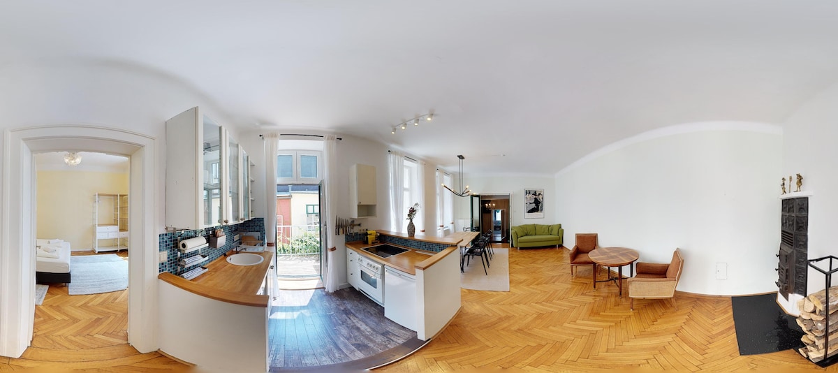 A spacious and bright interior is captured, showcasing a well-equipped kitchen with wooden countertops and a dining area. The living space features a comfortable sofa and a round table, while sunlight filters through large windows, illuminating the parquet flooring.