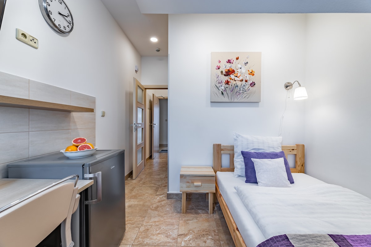 The bright, compact bedroom features a single bed with white bedding and decorative purple accents. A small bedside table and a wall-mounted light are visible. A kitchenette with a fridge and a small dining area is adjacent, with a clock adorning the wall.
