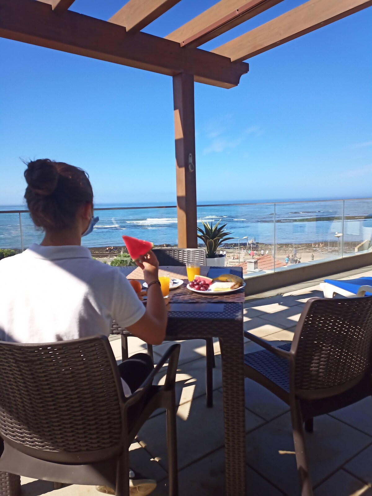 A dining area is positioned on a spacious balcony, featuring a table with breakfast items including fruit and drinks. A view of the ocean and beach can be seen in the background, framed by a wooden pergola and clear railing.