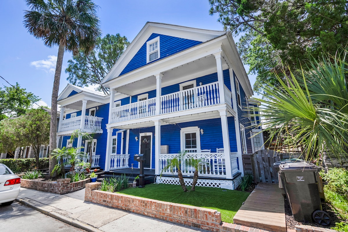A vibrant blue multi-unit building features two stories with white railings and large windows. Lush greenery and palm trees adorn the outdoor area, enhancing the charm of the property. The entrance is framed by neatly manicured grass and a brick walkway.