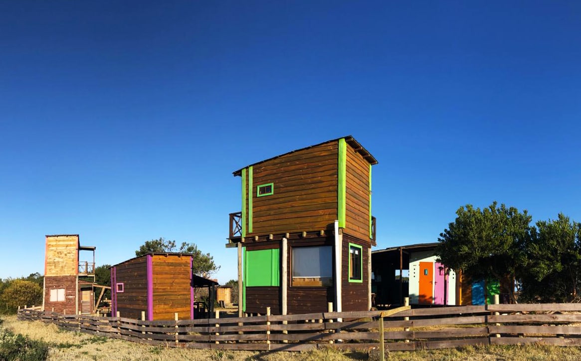 A collection of colorful mini cabañas is showcased against a clear blue sky. The wooden structures feature various vibrant door and window colors, complemented by a rustic wooden fence and surrounding greenery.