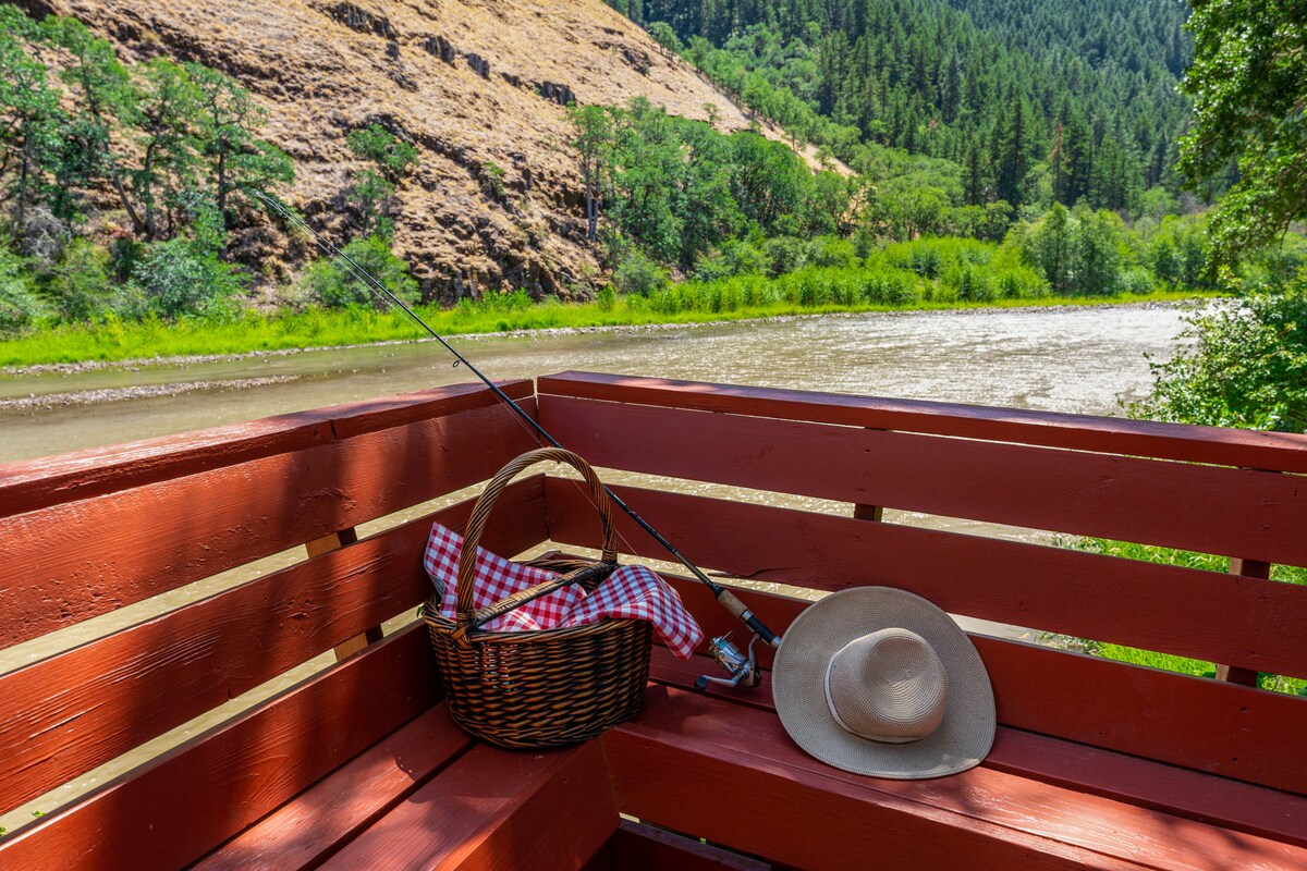 A red wooden bench is positioned along the riverbank, featuring a picnic basket lined with checkered cloth and a fishing rod resting nearby. Lush greenery and rolling hills are visible in the background, creating a serene outdoor setting by the river.