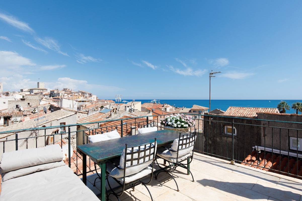 A spacious roof terrace features a large dining table surrounded by several chairs, offering panoramic views of the sea and rooftops of Antibes. The scene captures a bright blue sky, providing a serene backdrop for outdoor dining.