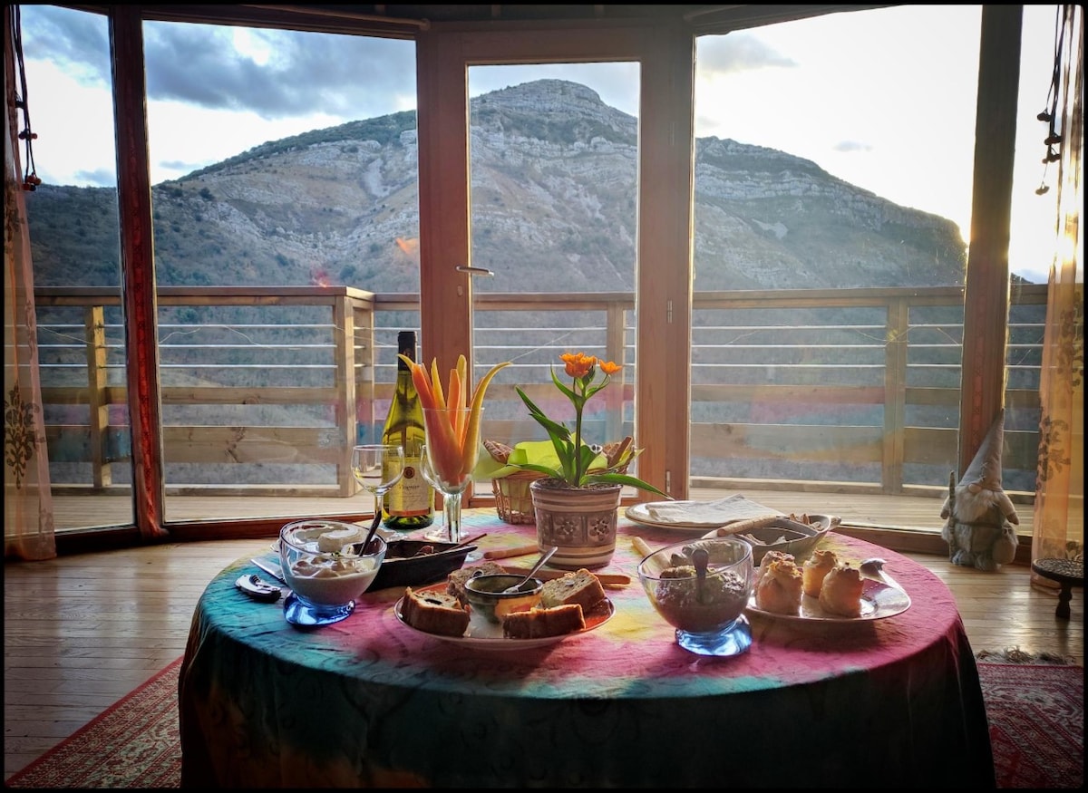 A round table is elegantly set with a variety of dishes and a vase containing flowers, while a bottle of champagne is displayed. Large glass doors overlook a mountainous landscape, allowing natural light to enhance the space. The floor is finished in natural wood.
