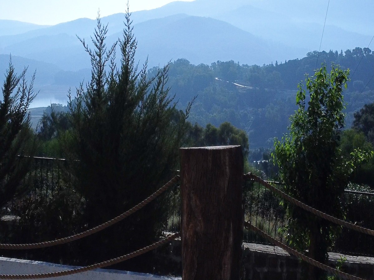 A panoramic view showcases layered mountains in soft blue hues, with lush greenery in the foreground. A sturdy wooden post stands surrounded by a natural rope barrier, offering a rustic touch to the serene landscape. The scene conveys a sense of peacefulness and connection to nature.