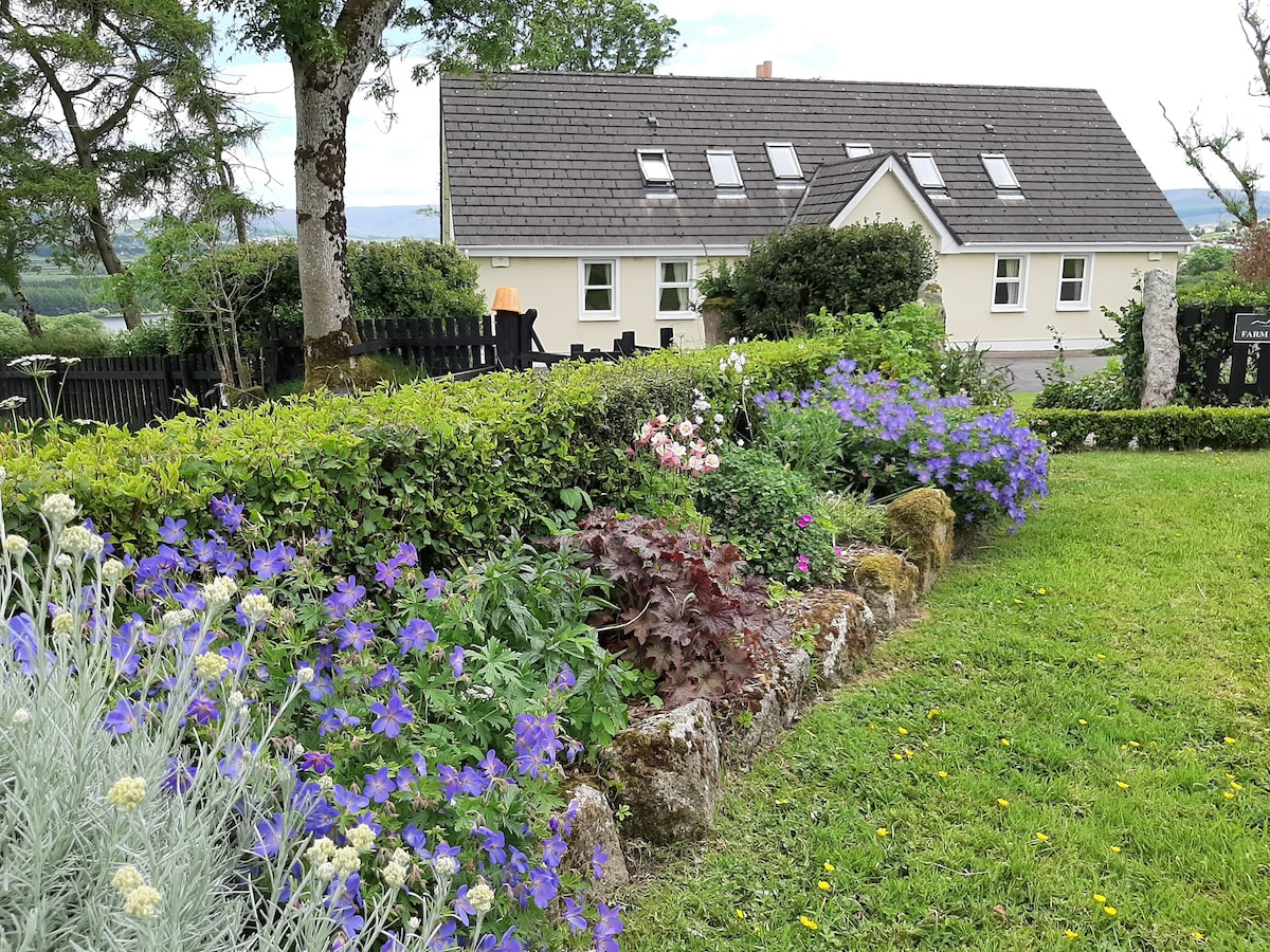 The image features a well-maintained garden with colorful flowers in various shades of purple, pink, and white. In the background, a spacious cottage with a slate roof and multiple skylights is visible, surrounded by lush greenery and neatly trimmed hedges.