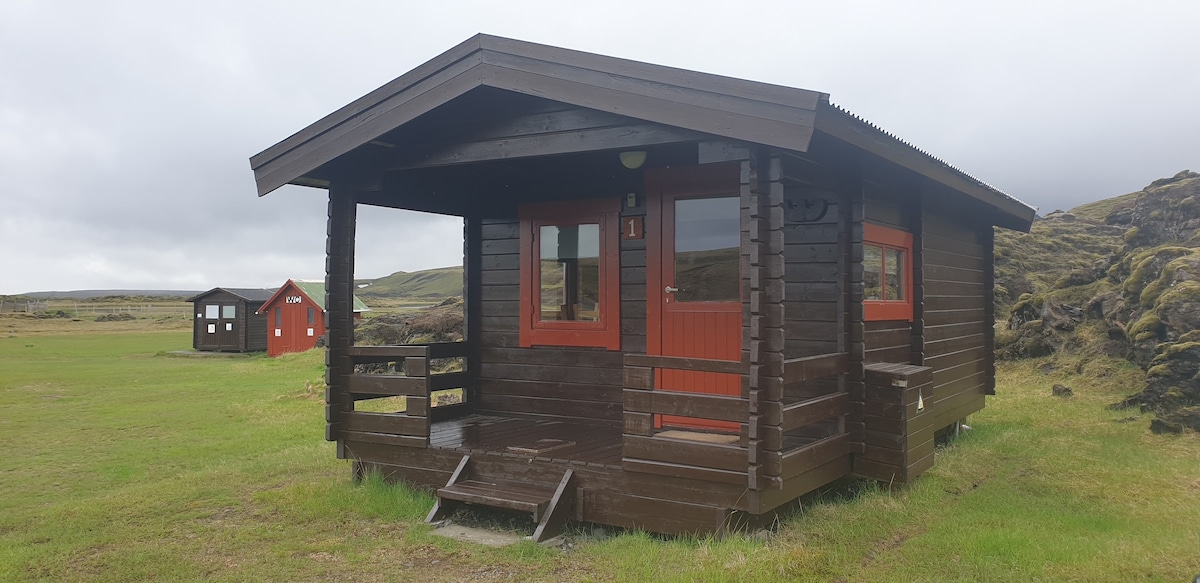A rustic wooden cabin features a slanted roof and bright red accents around the windows and doors. The entrance includes a small staircase leading up to a covered porch. Nearby, two other cabins can be seen in the lush green landscape, set against a backdrop of gentle hills.