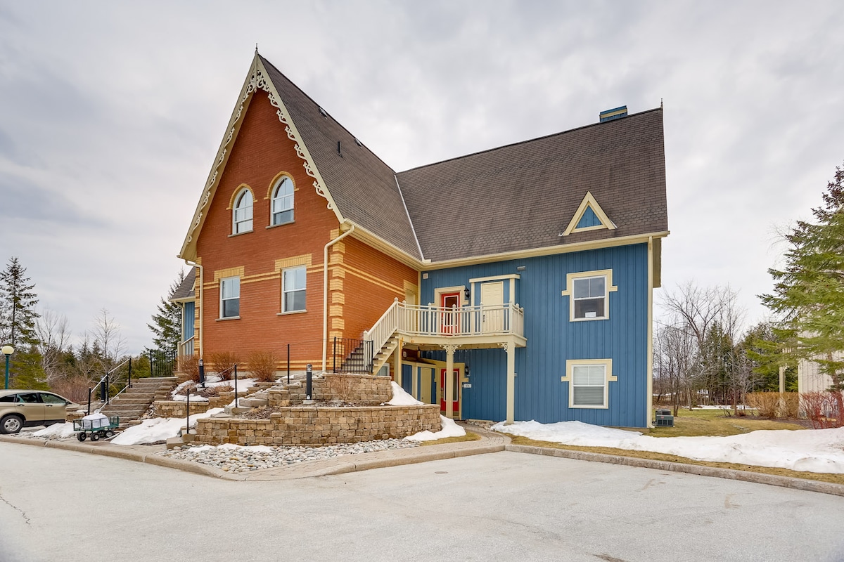 The exterior of a chalet-style condo is showcased, featuring a mix of red and blue siding with distinctive architectural details. A spacious front porch is accessible by stairs, and snow remains on the ground, indicating a winter setting. Nearby trees and the parking area complement the scene.