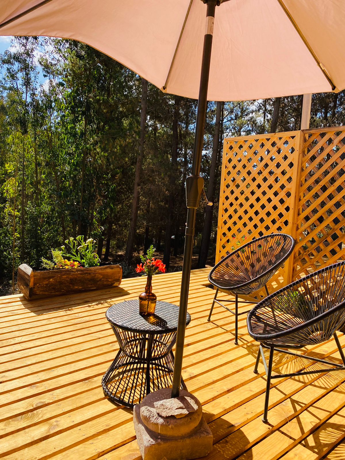 A wooden deck features two chairs and a small circular table under a beige umbrella. A planter with flowers is placed on one side, while a wooden privacy screen adds separation. The surrounding trees enhance the peaceful outdoor setting.