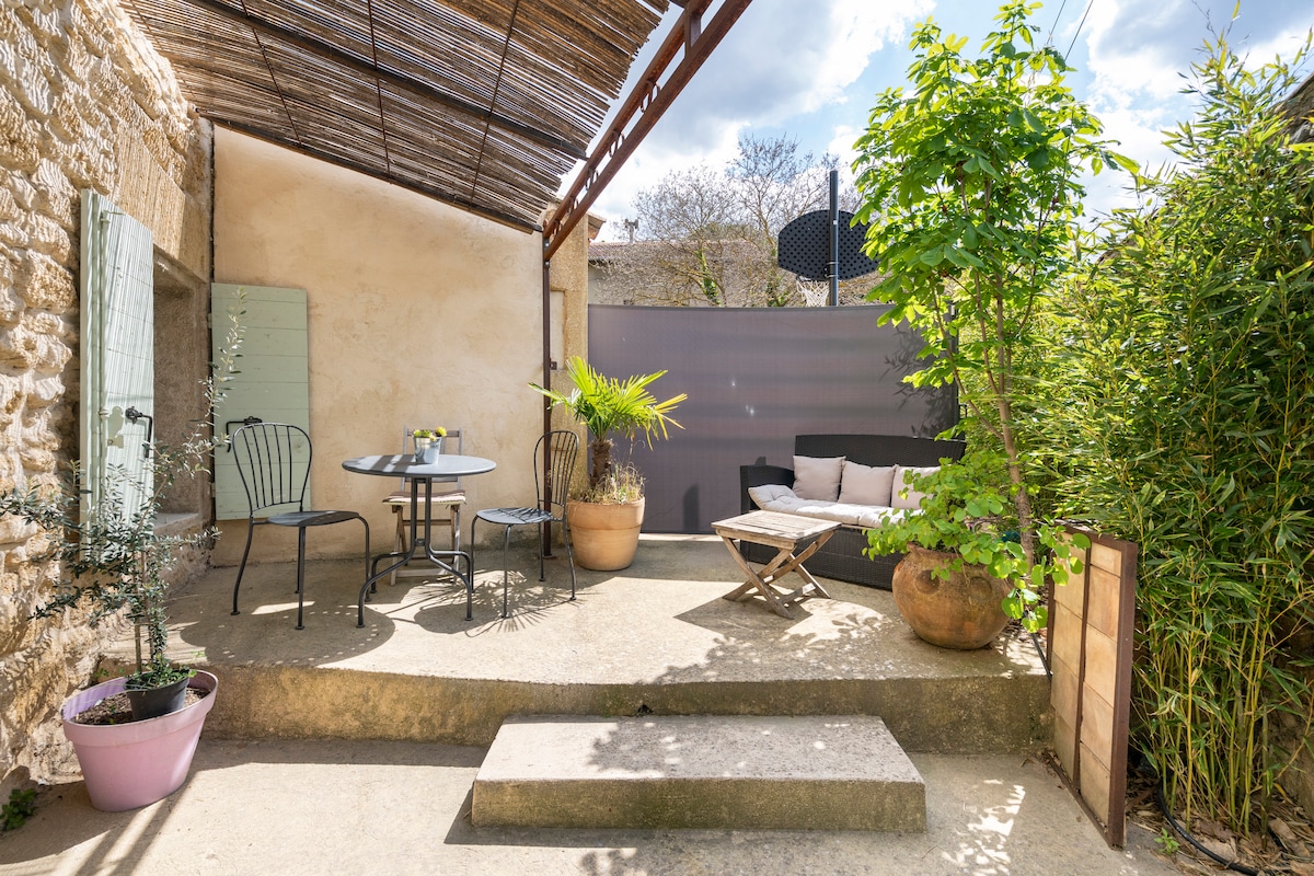An outdoor space features a small table with two chairs, surrounded by potted plants and greenery. A comfortable seating area offers a place to relax. The area is partially shaded, creating a cozy atmosphere for enjoying the sunshine.