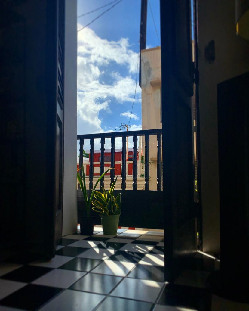 An entrance opens to a view of blue skies, with clouds visible from the doorway. A potted plant sits on the floor, surrounded by a patterned black and white floor that leads to the balcony adorned with wooden railings.