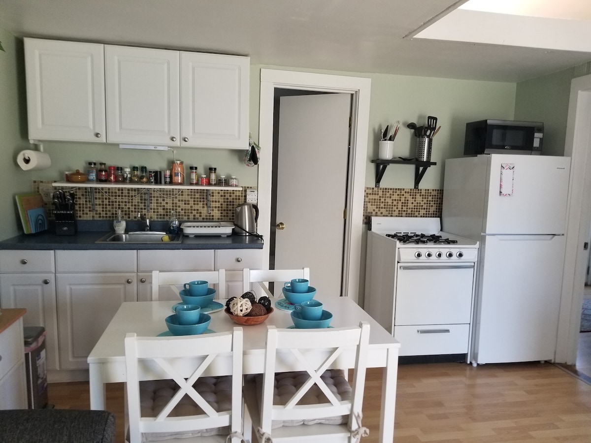 The kitchen area features a light green wall and white cabinetry, with a dining table set for four. A white stove, refrigerator, and microwave are present alongside neatly arranged kitchenware. Additional storage can be seen on the countertop, creating a functional cooking space.