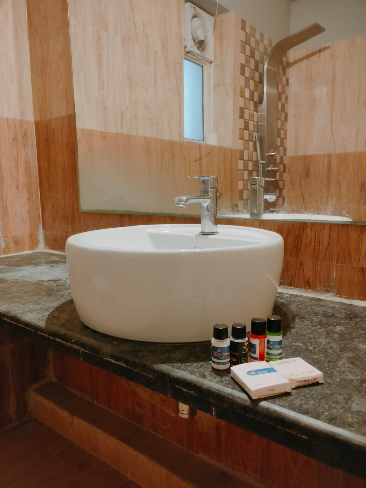 A modern bathroom features a round white basin set on a dark granite countertop. Several small toiletry items are neatly arranged beside the basin. A shower area with tiles can be seen in the background, enhanced by a mirror reflecting the space.