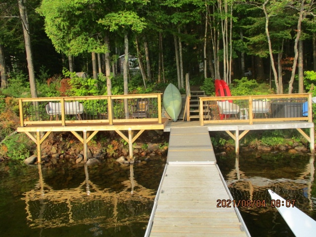 A wooden dock extends over the water, leading to a viewing deck surrounded by greenery. Comfortable seating is provided on the deck, which includes lounge chairs. A kayak is positioned beside the deck, offering a serene outdoor atmosphere.