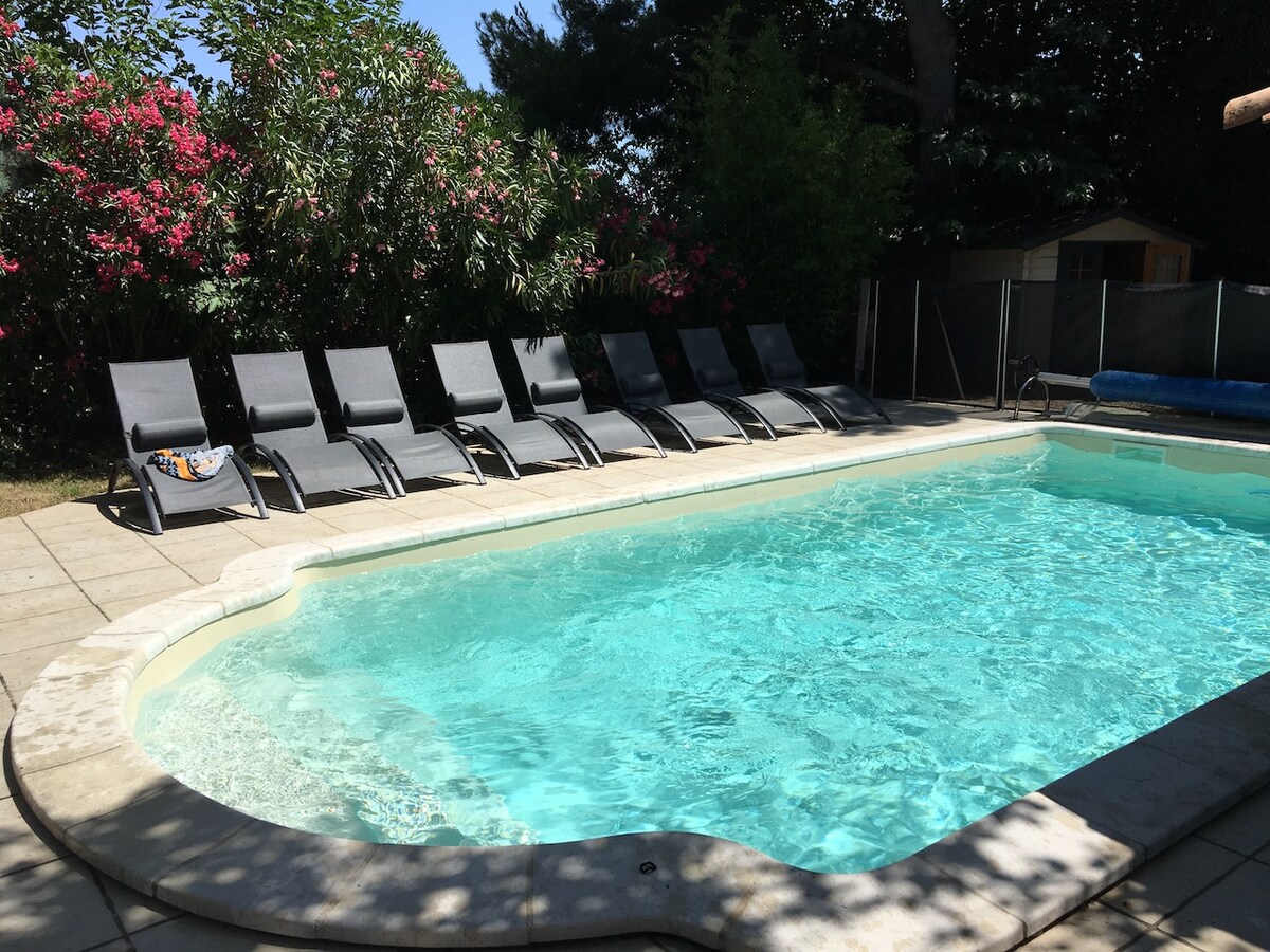 A private pool area features a sparkling blue pool surrounded by a tiled deck. Several lounge chairs are arranged neatly around the pool, offering space for relaxation. Lush greenery and flowering plants add a natural touch to the tranquil setting.
