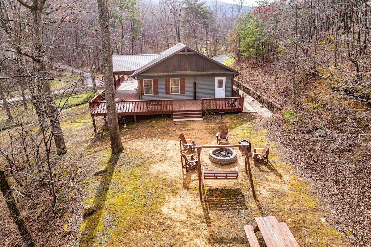 The cabin is surrounded by trees, showcasing a spacious outdoor area with a fire pit and seating arrangement. A picnic table is positioned nearby, and a large deck extends from the cabin, offering a welcoming space for relaxation amidst nature.