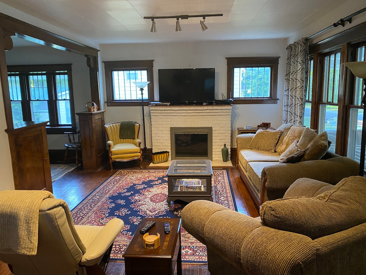 The living room features a spacious arrangement of comfortable seating, including a beige sectional and an armchair in soft yellow. A decorative area rug lays under a coffee table, with a television mounted above a white-brick fireplace. Natural light filters through multiple windows adorned with wooden frames.