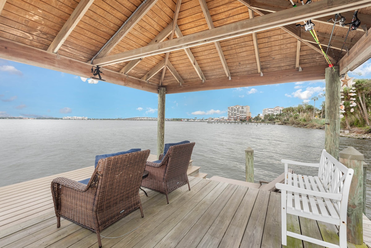 A wooden dock is seen extending over the water, offering a shaded area with a high wooden ceiling. Two patterned wicker chairs and a white bench provide seating, while the serene river view is complemented by distant buildings along the shore.