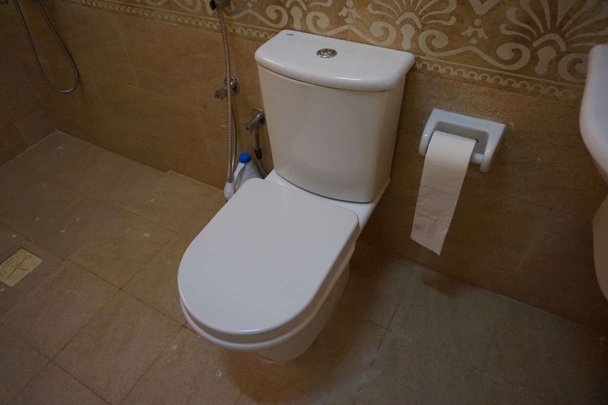 A clean and functional ensuite bathroom features a modern white toilet positioned on tiled flooring. Light beige tiles adorn the walls, offering a neutral backdrop. A roll of toilet paper is conveniently placed on a wall-mounted holder beside the toilet.