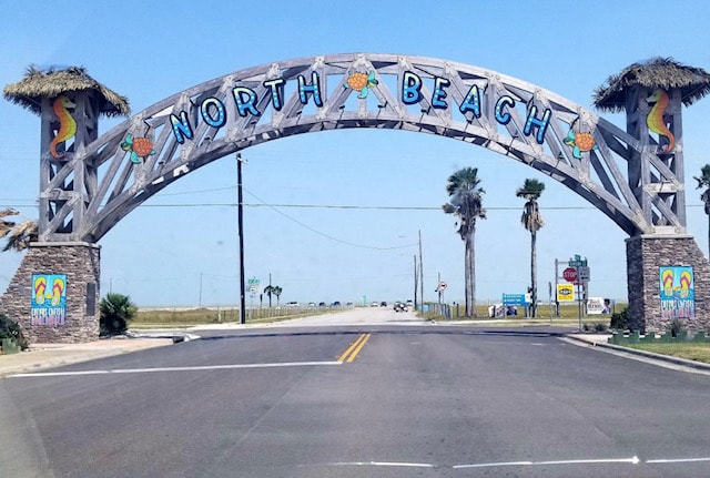 A large wooden archway marks the entrance to North Beach, featuring brightly colored lettering and palm trees. The scene is framed by a clear blue sky, emphasizing the coastal vibe of the area. A wide road leads into the beach, inviting visitors.