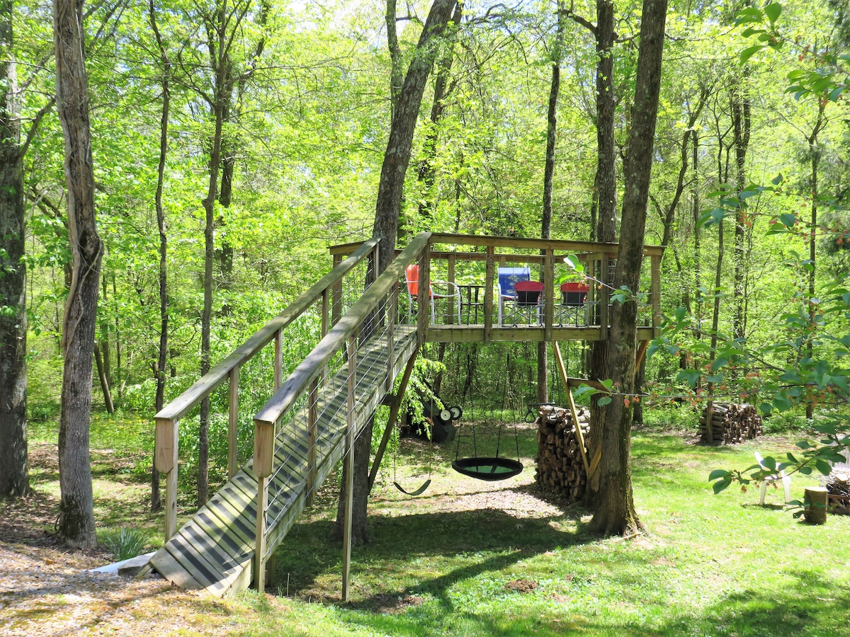 A wooden tree deck is elevated among vibrant green trees, accessed by a sloped walkway. Below, a swing hangs from a sturdy frame and firewood is stacked nearby. The surrounding area is spacious and grassy, creating an inviting outdoor environment.