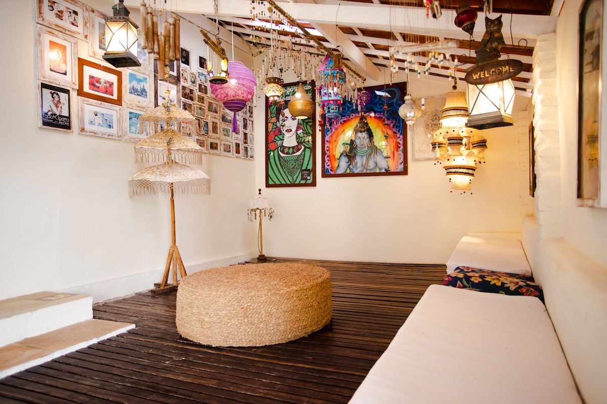 An inviting communal area features a circular woven seating arrangement at its center. Various decorative wall art pieces create a vibrant atmosphere, while multiple hanging lanterns and colorful accents enhance the space's character. Light wooden flooring adds warmth to this cozy environment.