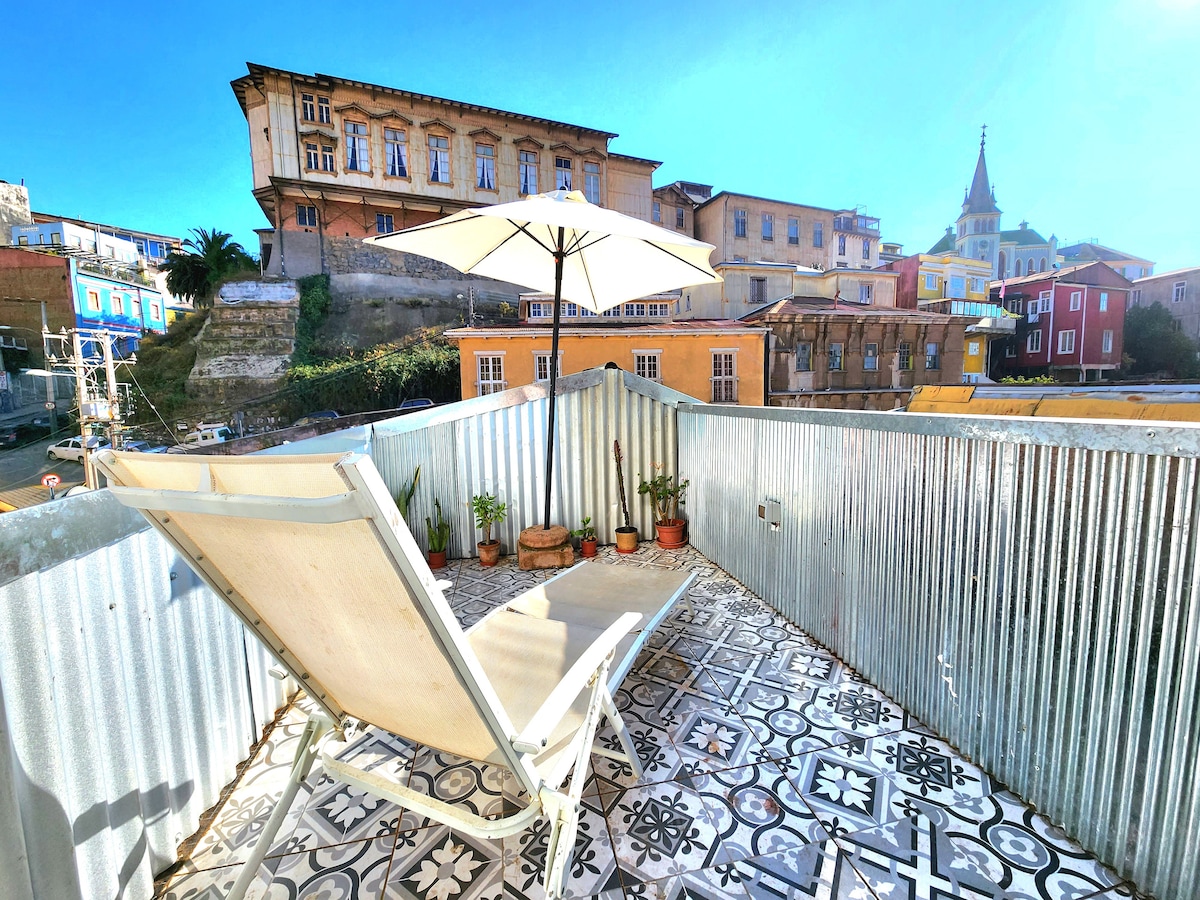 A rooftop terrace features a sun lounger positioned under a large umbrella. Decorative plants are placed in terracotta pots along the edge, with a view of historic buildings and a clear blue sky serving as a backdrop.