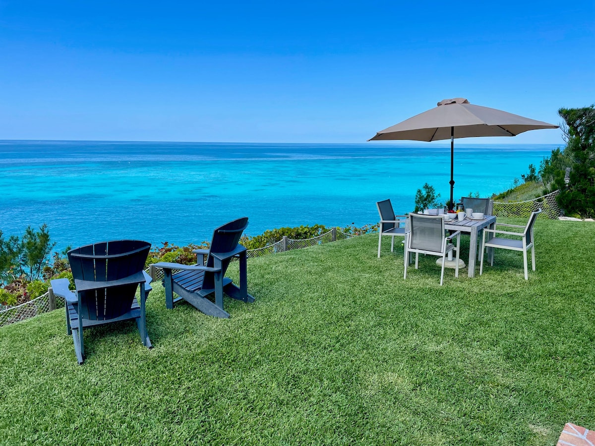 An outdoor seating area is set on a lush green lawn, featuring a large umbrella over a dining table surrounded by chairs. In the background, vibrant turquoise waters stretch toward a clear blue sky, creating a serene seaside atmosphere.