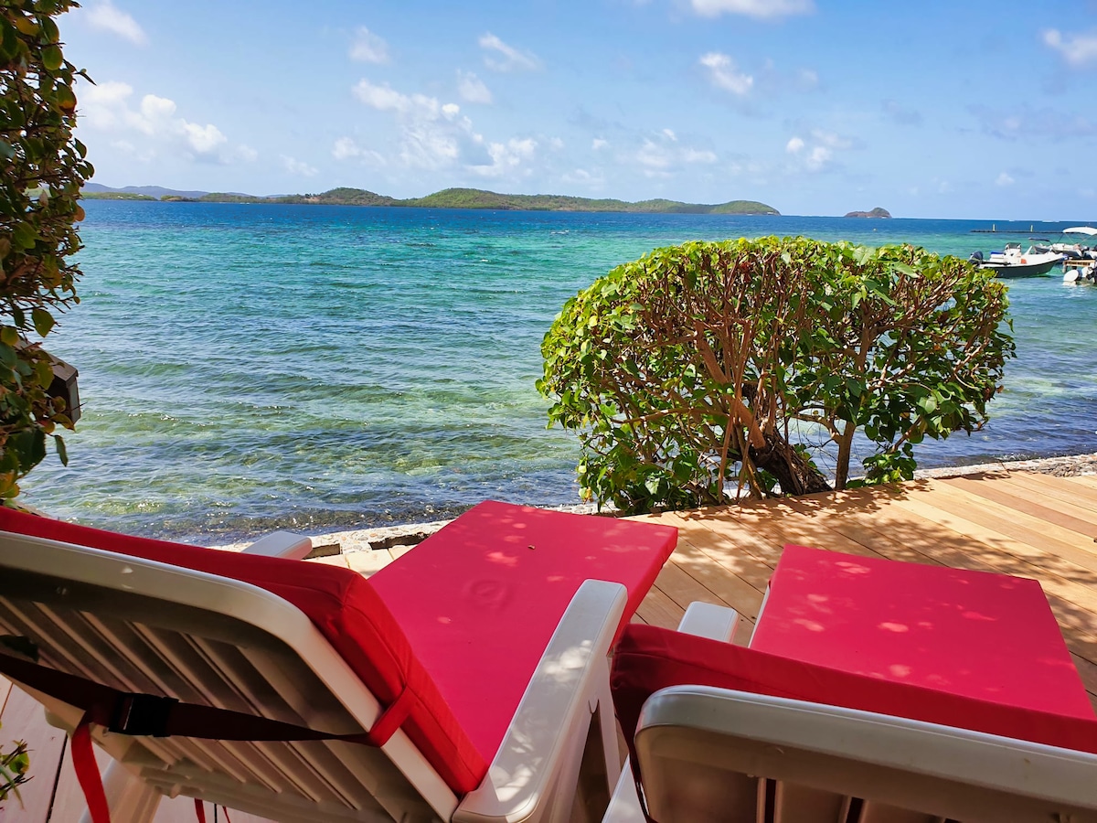 Two lounge chairs with red cushions are positioned on a wooden deck, facing a serene view of the ocean. Lush greenery features prominently in the foreground, while distant islands are visible across the turquoise waters under a clear blue sky.