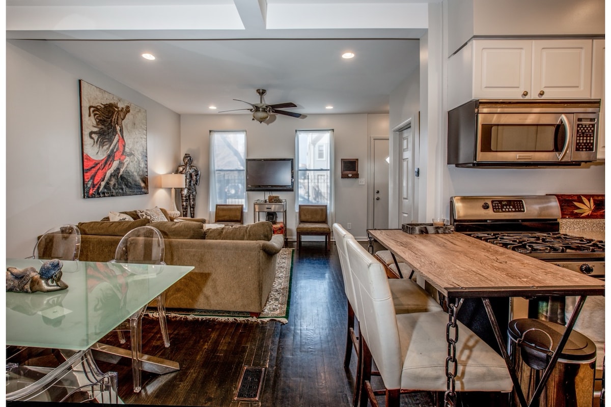 The living area showcases a comfortable seating arrangement with a large sofa and accent chairs. A modern kitchen is visible featuring stainless steel appliances and a dining table with transparent chairs. Natural light enters through multiple windows, enhancing the inviting atmosphere.